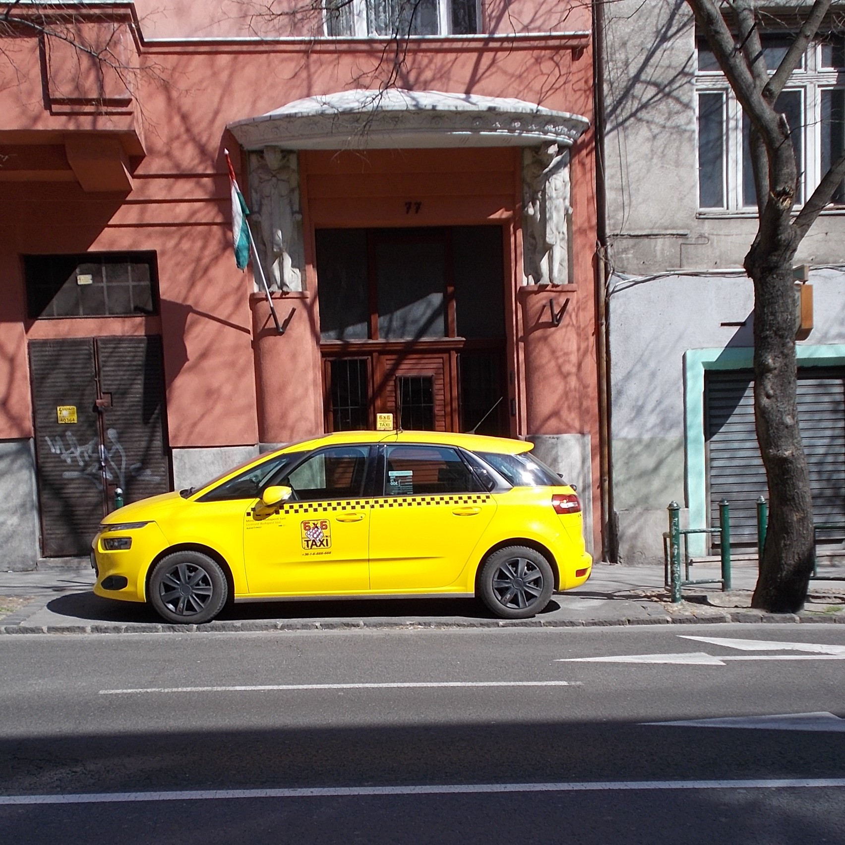 A yellow 6x6 Taxi Citroën parked on a Budapest street, with the firm's logo, phone number and chequered stripe on the doors
