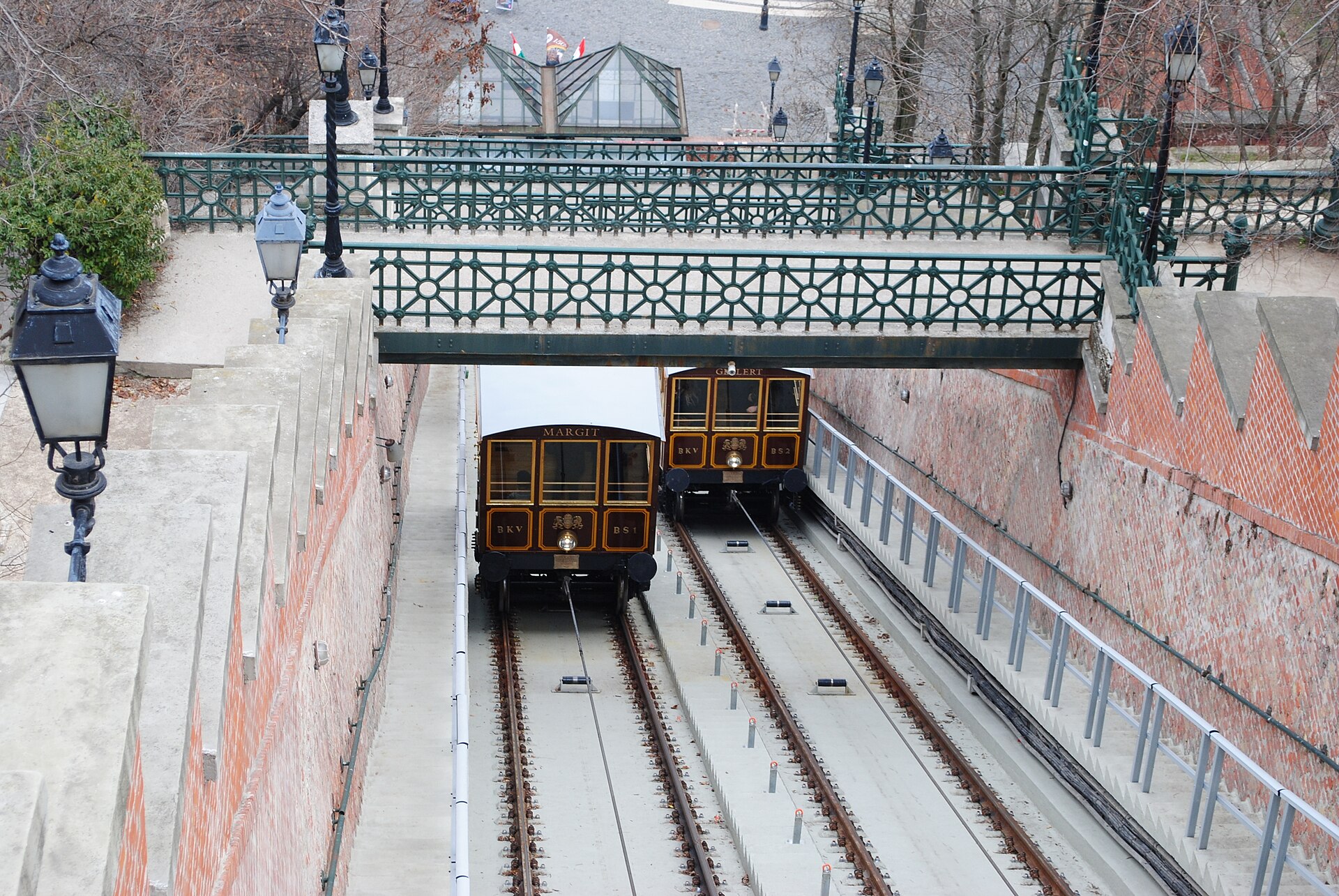 The two Buda Castle funicular cars named Margit and Gellert passing each other on the track below an ornate green iron bridge