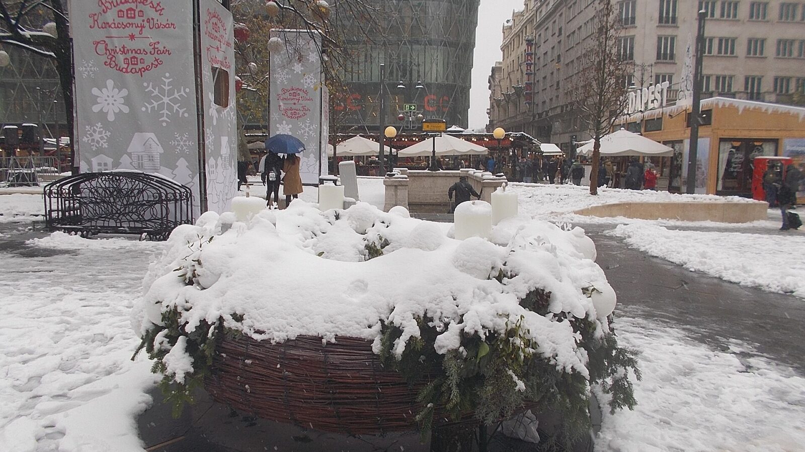 A giant Advent wreath with candles covered in snow on Vörösmarty tér, with the Budapest Christmas Fair signs and market stalls behind