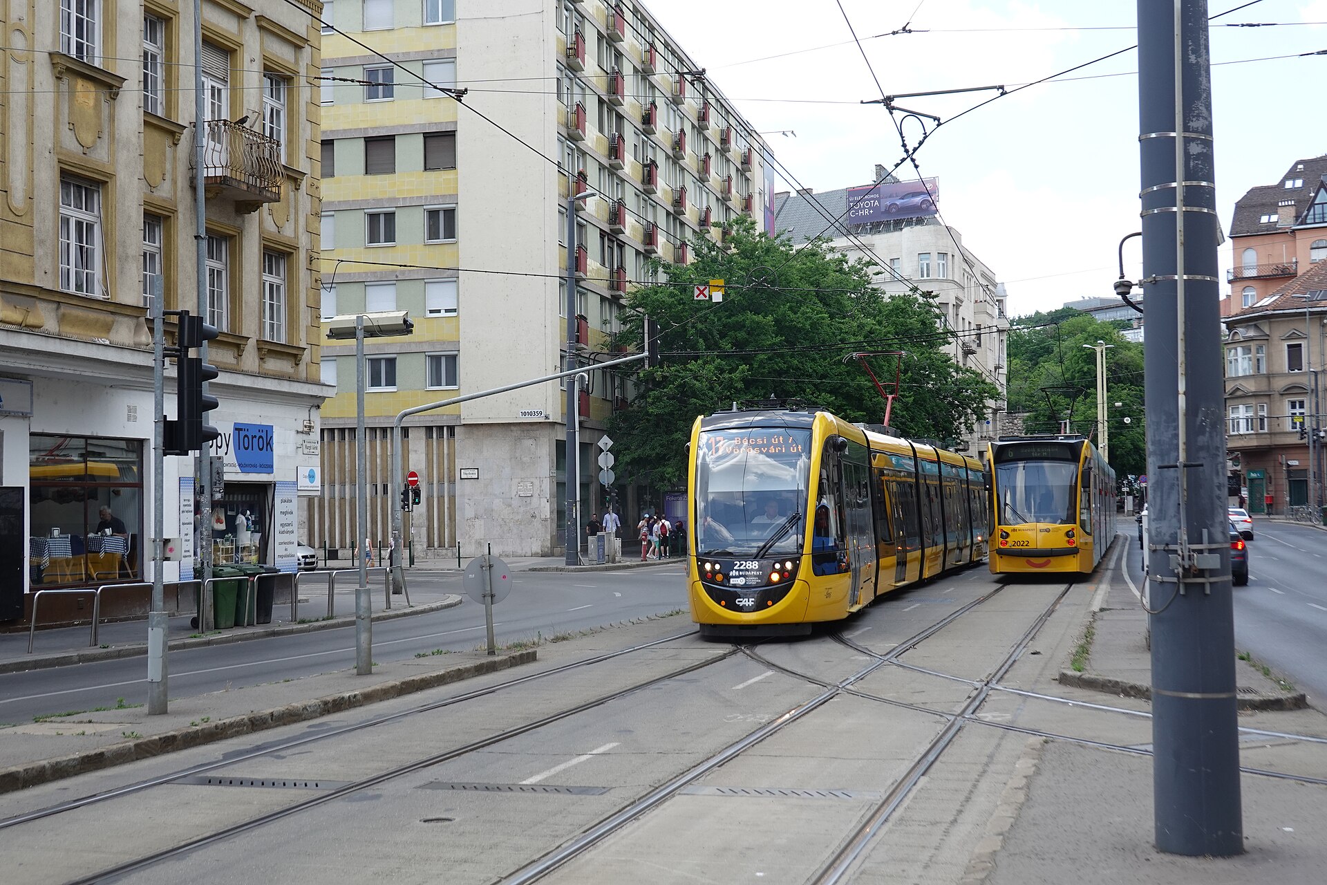 Two yellow CAF low-floor trams passing on a Buda street with overhead wires and apartment blocks behind