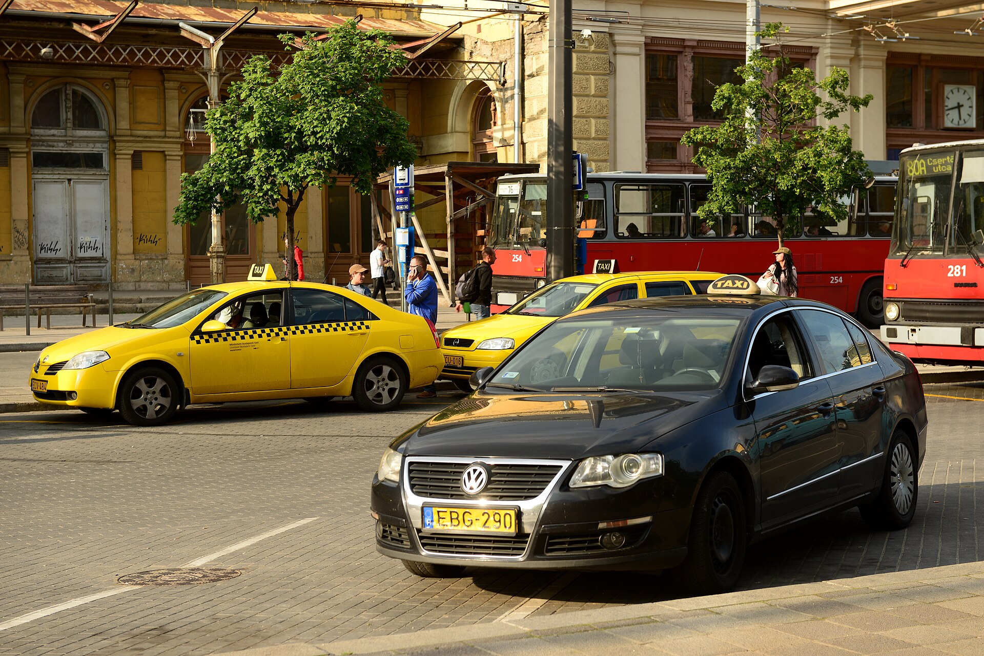 Yellow taxis waiting at a rank outside a Budapest railway station, with red trolleybuses behind