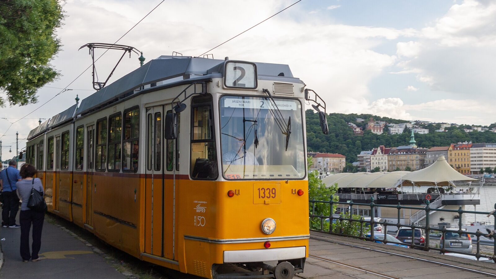 An orange-and-white tram on line 2, showing Jászai Mari tér – Közvágóhíd, on the Pest embankment with the Buda hills behind