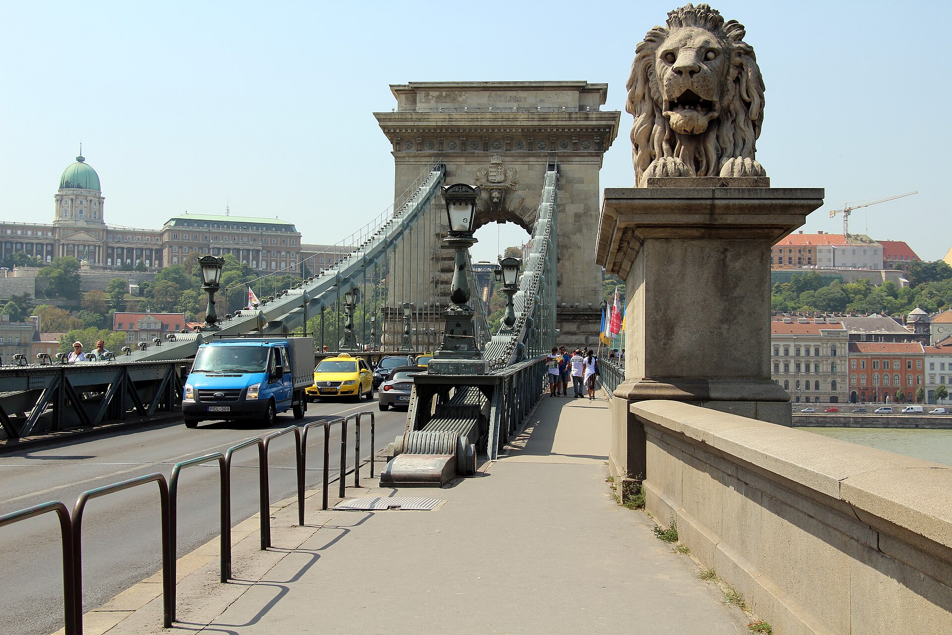 The Szechenyi Chain Bridge in Budapest from the pavement, with a stone lion on its plinth, the arched pylon beyond and Buda Castle on the hill behind