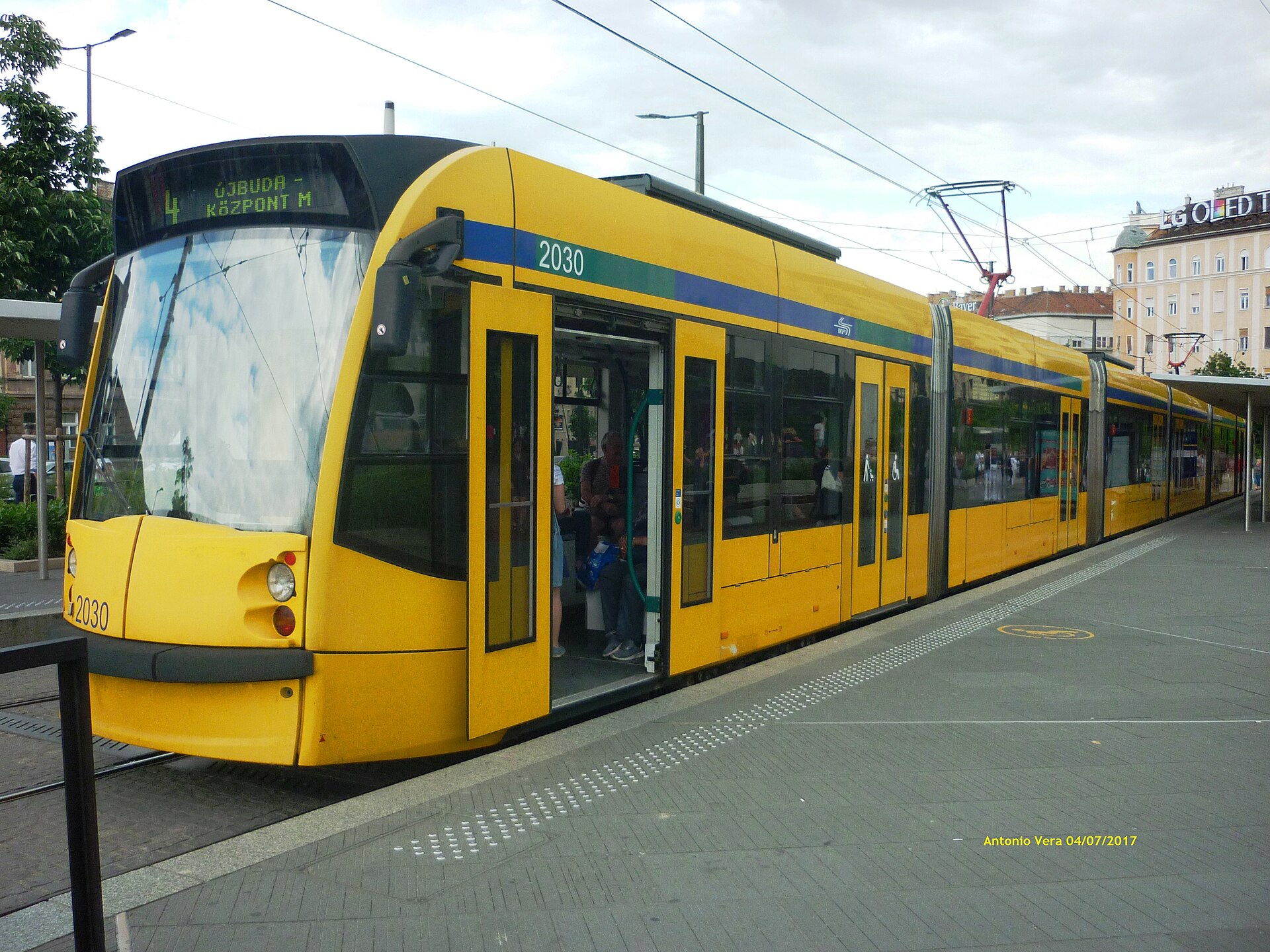 A long yellow Combino tram on line 4, bound for Újbuda-központ, with its doors open at a low-floor platform