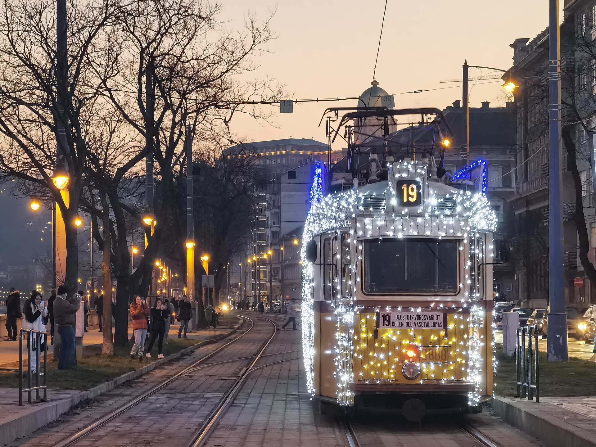 An old tram covered in thousands of white and yellow lights on line 19 along the Buda embankment at dusk, with the Royal Palace dome behind