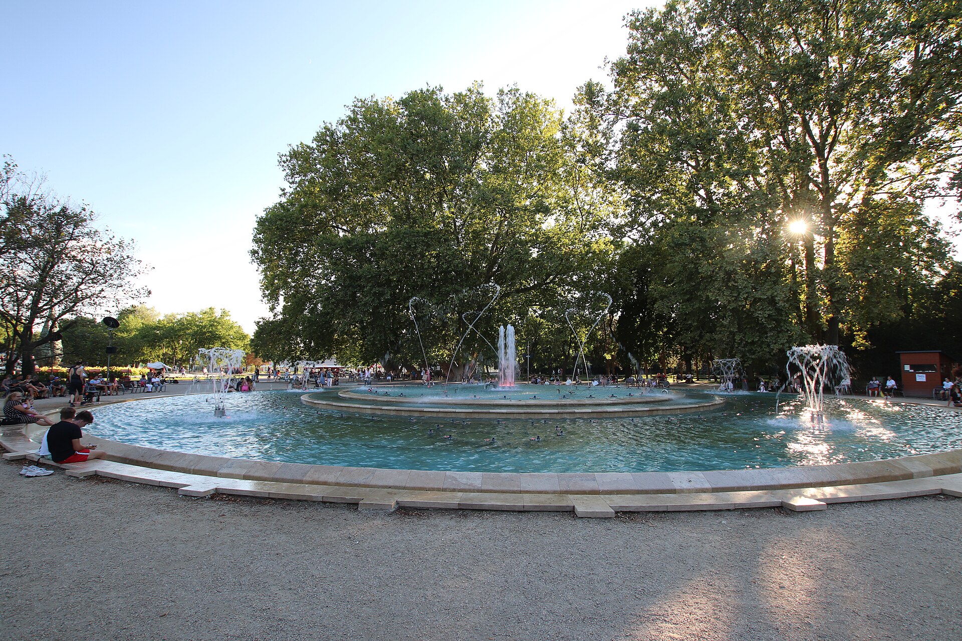 The large circular musical fountain on Margaret Island in Budapest with water jets playing and people sitting on the stone rim