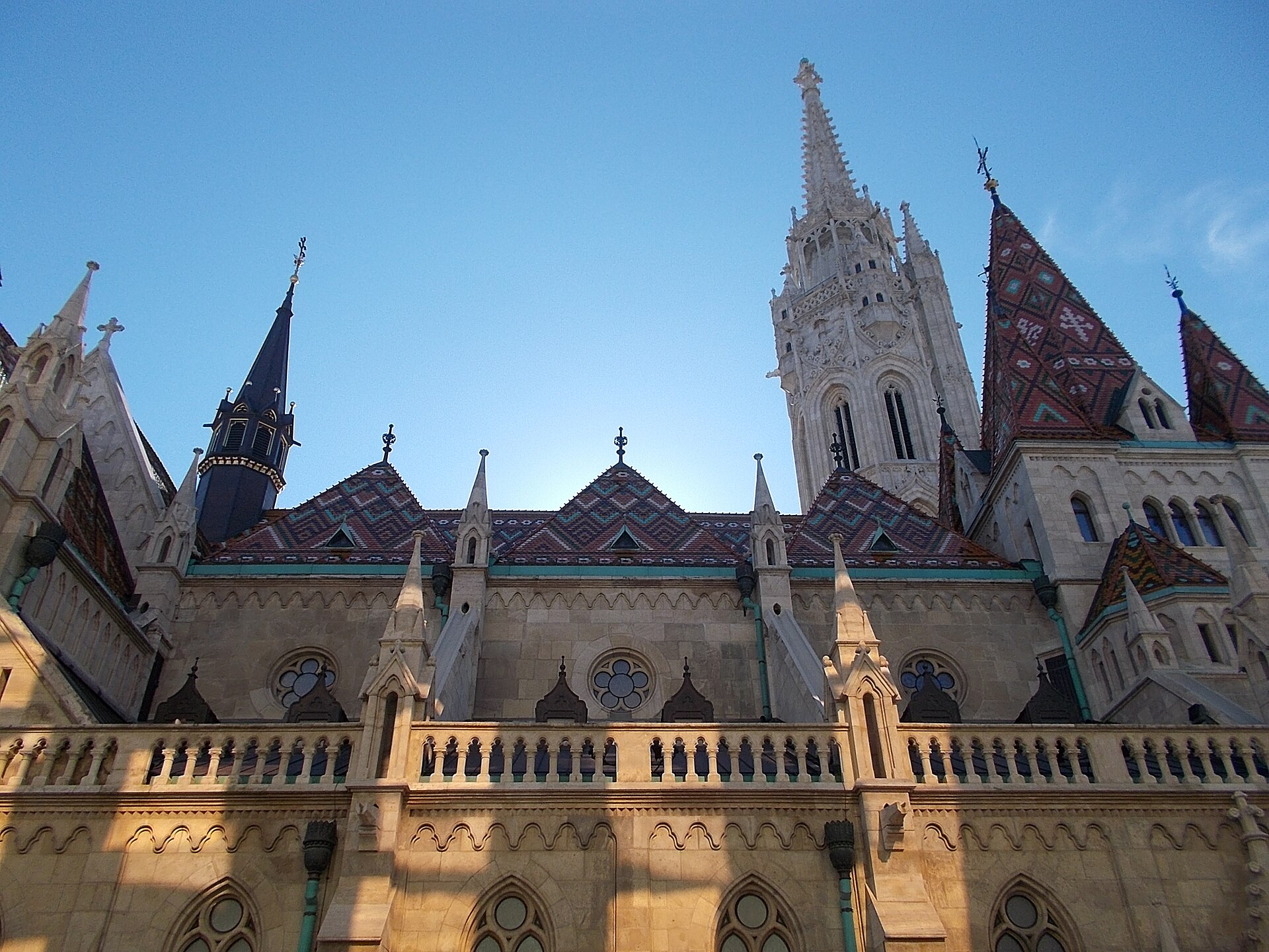 Matthias Church in Budapest seen from below in late afternoon light, showing the tiled roofs and the tall pierced stone Matthias Tower