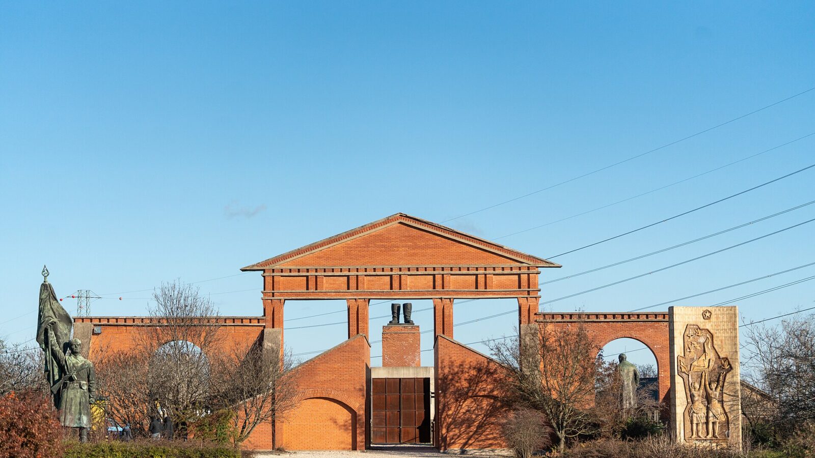 The brick entrance gate of Memento Park in Budapest, with statues flanking the sides and Stalin's boots monument visible through the columns