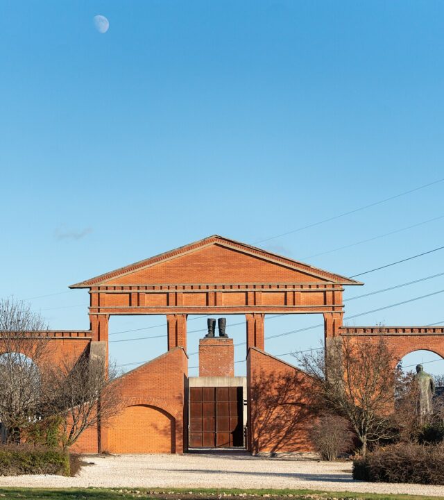 The brick entrance gate of Memento Park in Budapest, with statues flanking the sides and Stalin's boots monument visible through the columns