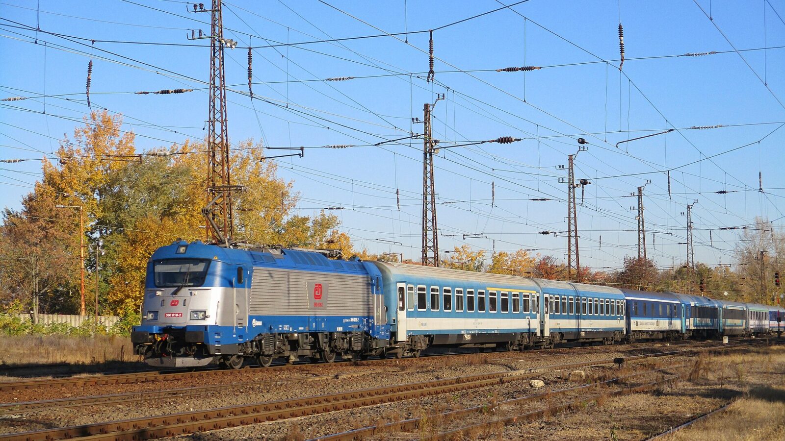 A Czech Railways electric locomotive hauling the Metropol night train's blue and white coaches and sleeping cars through a Hungarian station in autumn