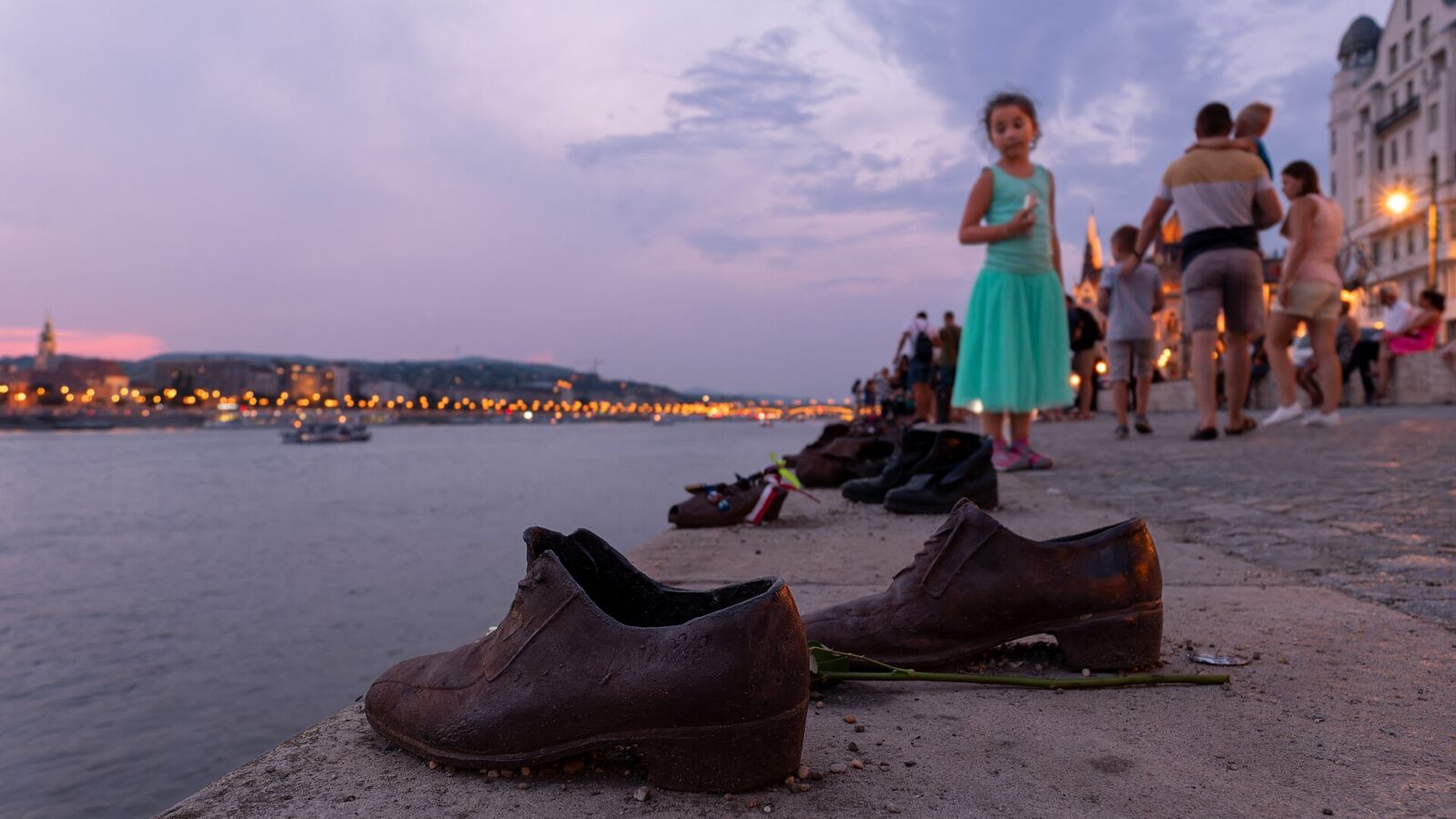 A pair of iron shoes at the edge of the Danube embankment at dusk with a rose laid beside them and the lit city behind