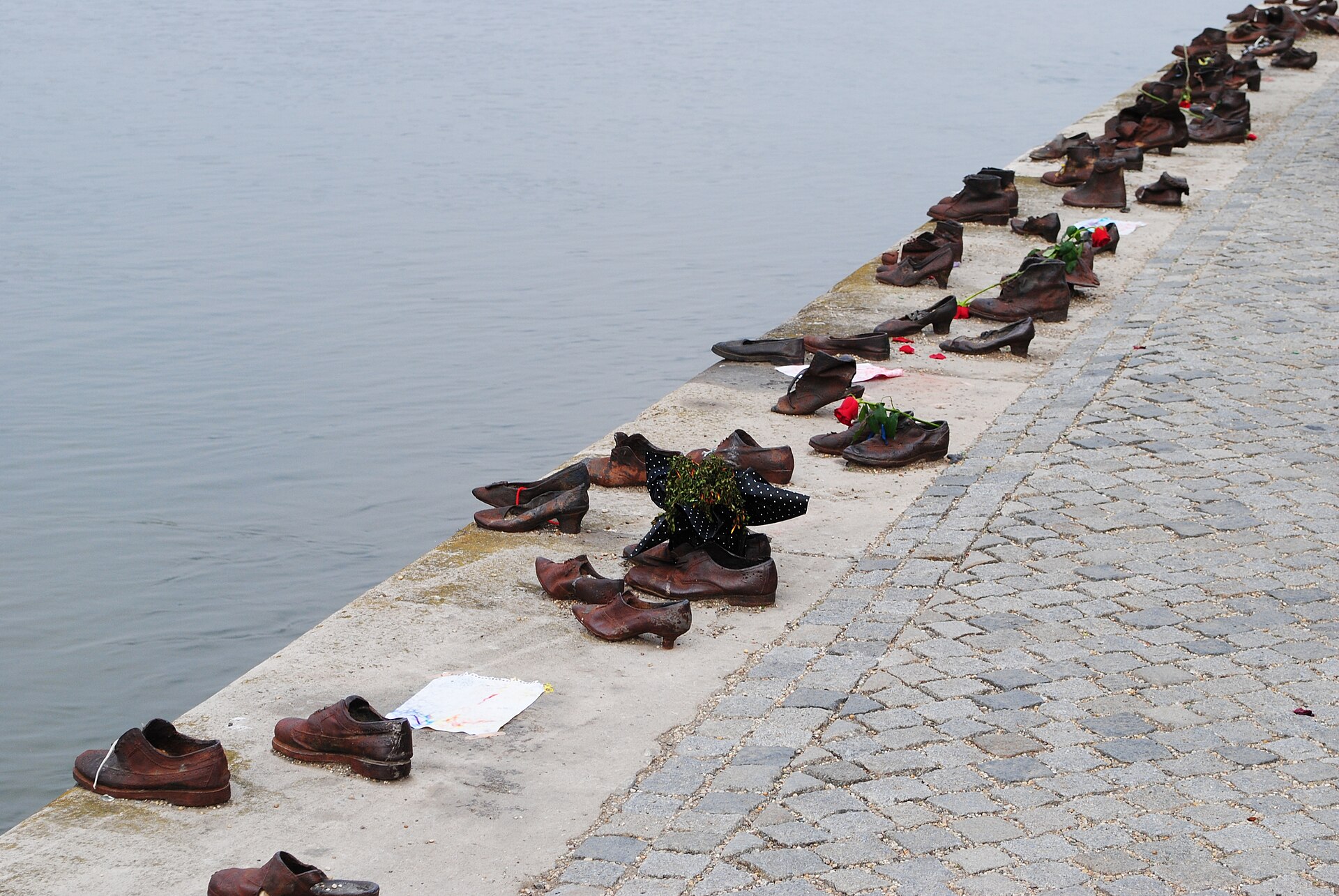 A line of rusted iron shoes fixed to the stone edge of the Danube embankment in Budapest, with flowers and a child's drawing left among them