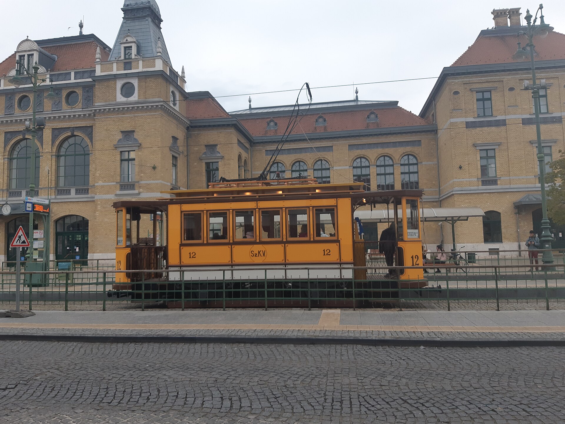 The yellow-brick facade of Szeged railway station, with a restored vintage tram painted yellow standing in front