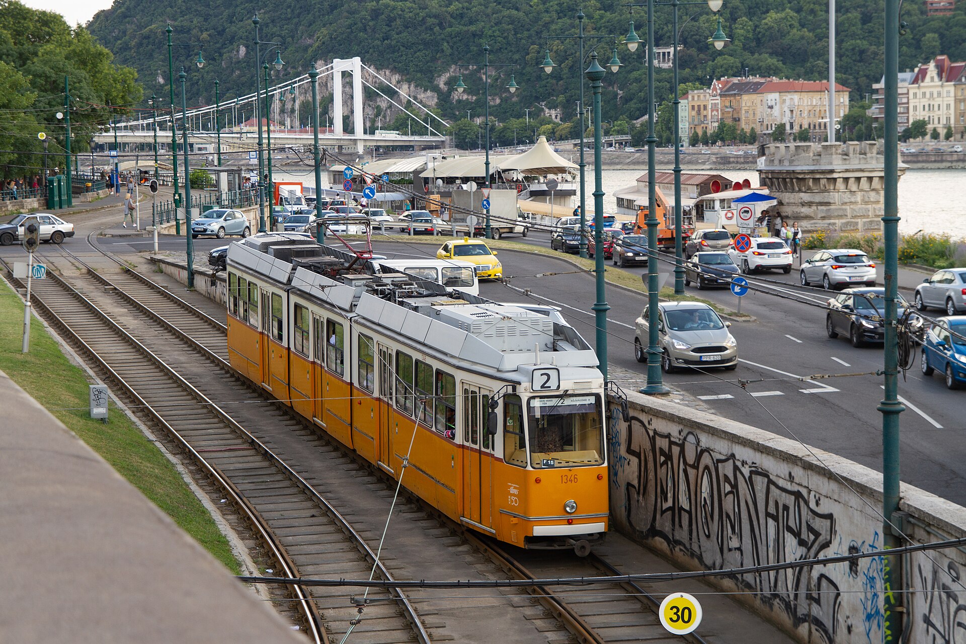An orange-and-white articulated tram on line 2 running beside the Danube on the Pest embankment, with the Elisabeth Bridge and Gellért Hill behind