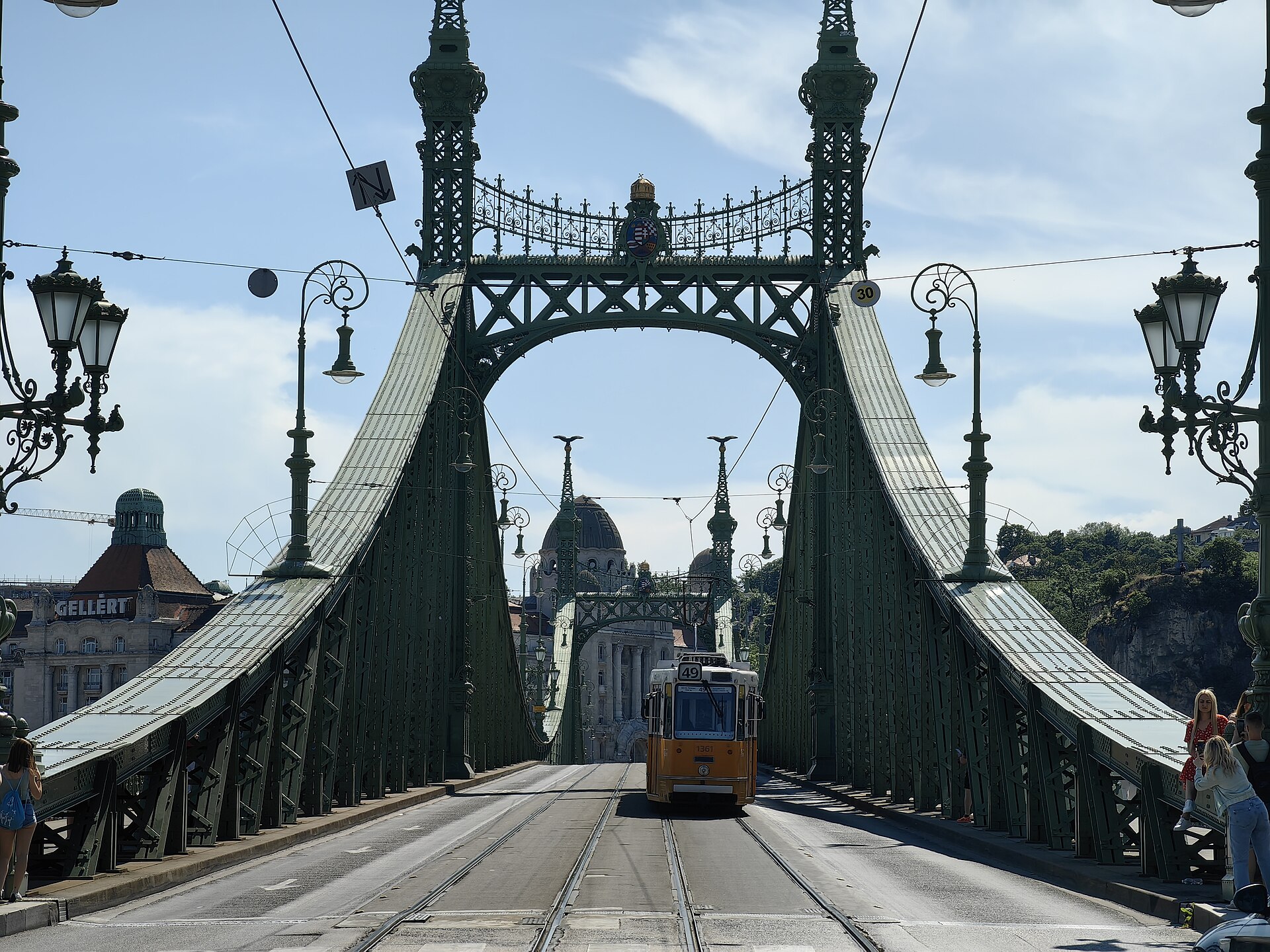 A yellow tram on line 49 crossing the green iron Liberty Bridge, with the Gellért hotel and baths at the Buda end